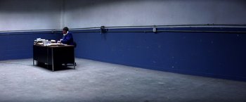 Movie still from “Punch-Drunk Love” (2002), directed by Paul Thomas Anderson – A person sitting in a chair in a room with a blue wall; Extreme Wide shot, Over the shoulder angle