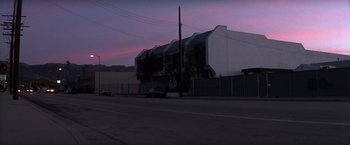 Movie still from “Punch-Drunk Love” (2002), directed by Paul Thomas Anderson – A car parked on the side of the road near a building; Extreme Wide shot, Low angle