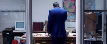 Movie still from “Punch-Drunk Love” (2002), directed by Paul Thomas Anderson – A man standing at a desk in front of a map of the united states; Wide shot, High angle