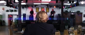 Movie still from “Punch-Drunk Love” (2002), directed by Paul Thomas Anderson – A man sitting in front of a stage with a band on it; Medium shot, Over the shoulder angle