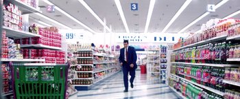 Movie still from “Punch-Drunk Love” (2002), directed by Paul Thomas Anderson – A man in a suit and tie walking in a grocery store aisle; Extreme Wide shot, High angle