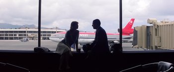 Movie still from “Punch-Drunk Love” (2002), directed by Paul Thomas Anderson – A man and a woman sitting on a bench in front of an airplane; Wide shot, Low angle