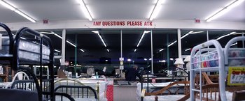 Movie still from “Punch-Drunk Love” (2002), directed by Paul Thomas Anderson – A man standing in front of a room with a sign on the wall; Extreme Wide shot, High angle