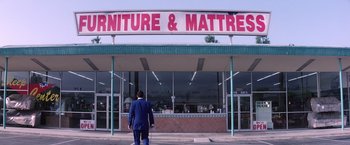 Movie still from “Punch-Drunk Love” (2002), directed by Paul Thomas Anderson – A man standing in front of a furniture and mattress store; Extreme Wide shot, Low angle