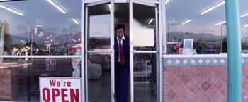 Movie still from “Punch-Drunk Love” (2002), directed by Paul Thomas Anderson – A man in a blue suit and red tie standing in front of an open door; Wide shot, Low angle