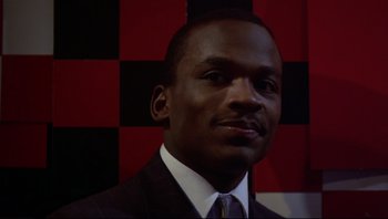 Movie still from “Purple Rain” (1984), directed by Albert Magnoli – A man wearing a suit and tie in front of a checkerboard wall; Close Up shot, Low angle