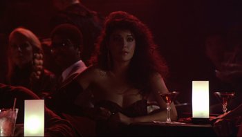 Movie still from “Purple Rain” (1984), directed by Albert Magnoli – A woman sitting at a table with a glass in front of her; Medium shot, Over the shoulder angle