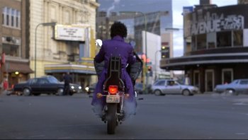 Movie still from “Purple Rain” (1984), directed by Albert Magnoli – A person riding a motorcycle down a street; Wide shot, Low angle