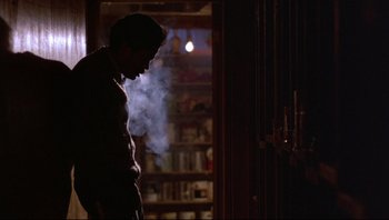 Movie still from “Purple Rain” (1984), directed by Albert Magnoli – A man standing in front of a bookshelf with smoke coming out of his mouth; Wide shot, Low angle