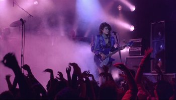 Movie still from “Purple Rain” (1984), directed by Albert Magnoli – A person on a stage playing a guitar; Wide shot, Low angle
