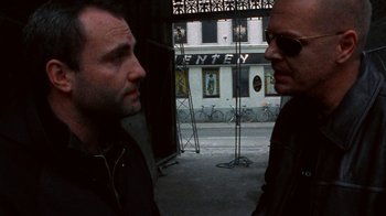 Movie still from “Pusher” (1996), directed by Nicolas Winding Refn – Two men standing next to each other in front of a building; Medium shot, Low angle