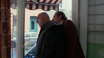 Movie still from “Pusher” (1996), directed by Nicolas Winding Refn – Two men standing next to each other in front of a building; Medium shot, Low angle