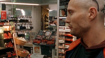 Movie still from “Pusher” (1996), directed by Nicolas Winding Refn – A man standing in front of a display case of cosmetics; Close Up shot, Over the shoulder angle