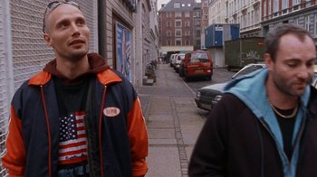 Movie still from “Pusher” (1996), directed by Nicolas Winding Refn – A man standing on the side of a street; Medium shot, Low angle