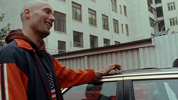 Movie still from “Pusher” (1996), directed by Nicolas Winding Refn – A man standing next to a car in a parking lot; Medium shot, Low angle