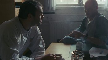 Movie still from “Pusher” (1996), directed by Nicolas Winding Refn – A man sitting at a table with a cup of coffee; Medium shot, Over the shoulder angle