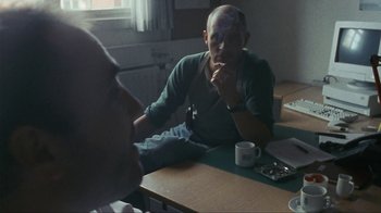 Movie still from “Pusher” (1996), directed by Nicolas Winding Refn – A man sitting at a wooden table with a cup of coffee; Medium shot, Over the shoulder angle
