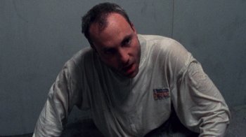 Movie still from “Pusher” (1996), directed by Nicolas Winding Refn – A man in a white t - shirt is sitting down; Close Up shot, Overhead angle