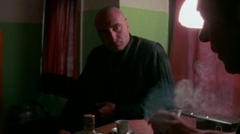 Movie still from “Pusher” (1996), directed by Nicolas Winding Refn – A man sitting at a table with a cigarette in his mouth; Medium shot, Over the shoulder angle