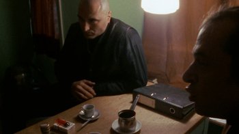 Movie still from “Pusher” (1996), directed by Nicolas Winding Refn – A man sitting at a table with a cup of coffee; Medium shot, Over the shoulder angle