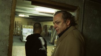 Movie still from “Pusher III” (2005), directed by Nicolas Winding Refn – A man standing next to another man in a warehouse; Medium shot, Over the shoulder angle