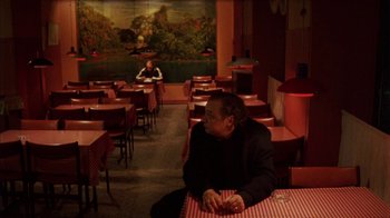 Movie still from “Pusher III” (2005), directed by Nicolas Winding Refn – A man sitting at a table in front of a painting; Wide shot, High angle