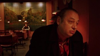 Movie still from “Pusher III” (2005), directed by Nicolas Winding Refn – A man sitting at a table in front of a painting; Close Up shot, Low angle