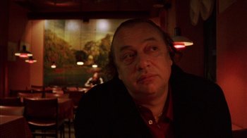 Movie still from “Pusher III” (2005), directed by Nicolas Winding Refn – A man in a red shirt is sitting in front of a painting; Close Up shot, Low angle