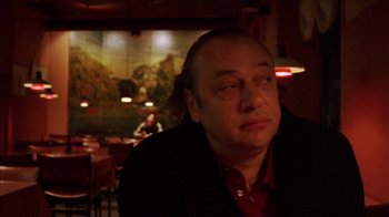 Movie still from “Pusher III” (2005), directed by Nicolas Winding Refn – A man sitting at a table in front of a painting; Close Up shot, Low angle