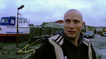 Movie still from “Pusher II” (2004), directed by Nicolas Winding Refn – A man with a beard is standing in front of a boat; Close Up shot, Low angle