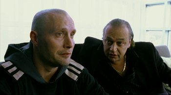 Movie still from “Pusher II” (2004), directed by Nicolas Winding Refn – Two men sitting next to each other in front of a wall; Close Up shot, Over the shoulder angle