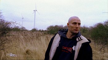 Movie still from “Pusher II” (2004), directed by Nicolas Winding Refn – A man standing in a field with a wind turbine in the background; Close Up shot, Low angle