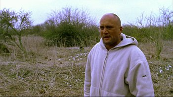 Movie still from “Pusher II” (2004), directed by Nicolas Winding Refn – A bald man in a white jacket standing in a field; Medium shot, Low angle
