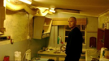 Movie still from “Pusher II” (2004), directed by Nicolas Winding Refn – A man standing in a kitchen next to a sink; Medium shot, Low angle
