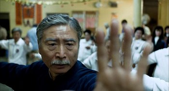 Movie still from “Pushing Hands” (1991), directed by Ang Lee – An older man holding up his hand in front of a group of people; Close Up shot, Over the shoulder angle