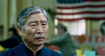 Movie still from “Pushing Hands” (1991), directed by Ang Lee – An older man in a blue shirt with a flag in the background; Close Up shot, Over the shoulder angle