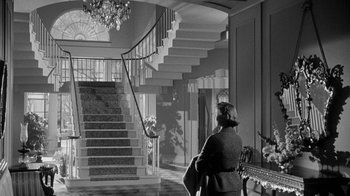 Movie still from “Queen Bee” (1955), directed by Ranald MacDougall – An old photo of a woman sitting in front of a staircase; Wide shot, Low angle