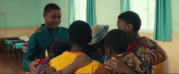 Movie still from “Queen of Katwe” (2016), directed by Mira Nair – A group of people standing around each other in a room; Medium shot, Over the shoulder angle