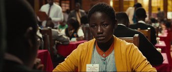 Movie still from “Queen of Katwe” (2016), directed by Mira Nair – A woman in a yellow jacket is sitting in front of a group of people; Close Up shot, Over the shoulder angle