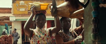 Movie still from “Queen of Katwe” (2016), directed by Mira Nair – Two young women are standing together and one of them is holding a basket on top of her head; Medium shot, Low angle