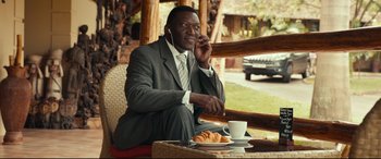 Movie still from “Queen of Katwe” (2016), directed by Mira Nair – A man in a suit and tie sitting in a chair with a cup of coffee and croissant; Medium shot, Over the shoulder angle