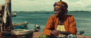Movie still from “Queen of Katwe” (2016), directed by Mira Nair – A woman sitting on the beach looking out at the water; Medium shot, Over the shoulder angle