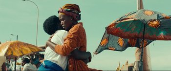 Movie still from “Queen of Katwe” (2016), directed by Mira Nair – Two women embracing each other in front of an umbrella; Medium shot, Low angle