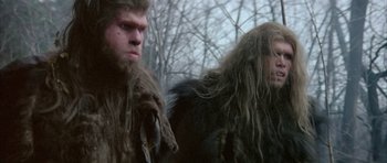 Movie still from “Quest for Fire” (1981), directed by Jean-Jacques Annaud – A man with long hair and a long beard is standing next to another man with long hair and a long beard; Close Up shot, Low angle