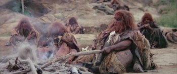 Movie still from “Quest for Fire” (1981), directed by Jean-Jacques Annaud – A group of people dressed as cavemen are gathered around a fire; Wide shot, High angle