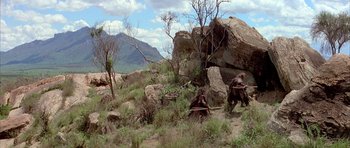 Movie still from “Quest for Fire” (1981), directed by Jean-Jacques Annaud – Two people are walking through the grass near rocks and trees; Extreme Wide shot, High angle