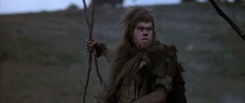 Movie still from “Quest for Fire” (1981), directed by Jean-Jacques Annaud – A man with a long beard holding a stick in a field; Medium shot, Low angle