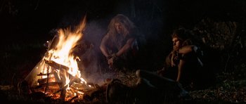 Movie still from “Quest for Fire” (1981), directed by Jean-Jacques Annaud – A woman sitting on the ground near a fire; Wide shot, Low angle