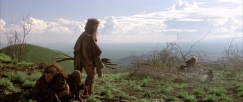 Movie still from “Quest for Fire” (1981), directed by Jean-Jacques Annaud – A man in a costume standing on top of a grass covered hill; Wide shot, Low angle