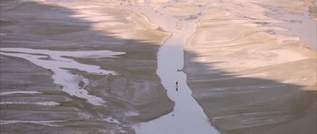 Movie still from “Quest for Fire” (1981), directed by Jean-Jacques Annaud – A person is walking on the beach near the water; Extreme Wide shot, High angle